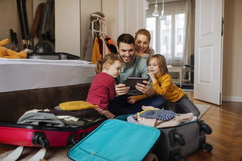 Junge Familie schaut auf ein Tablet während Sie vor gepackten Koffern in ihrer Wohnung sitzen. 