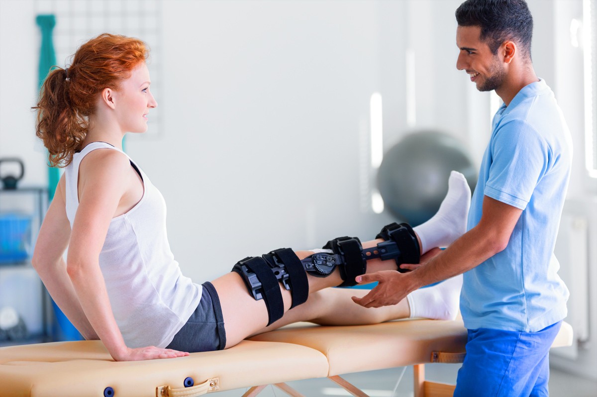 Ein circa 17-jähriges Mädchen mit Schiene am rechten Bein sitzt beim Physiotherapeuten auf der Liege. Er hebt ihr Bein an.