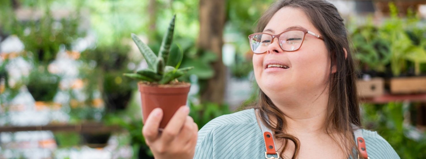 Eine etwa 20 jährige Frau mit Down-Syndrom hält eine Topflanze in der rechten Hand.