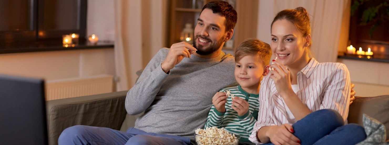 Eine Familie sitzt gemeinsam auf der Couch und schaut interessiert zum Fernseher. Der Junge hat eine Popcornschale auf dem Schoß und alle naschen davon.