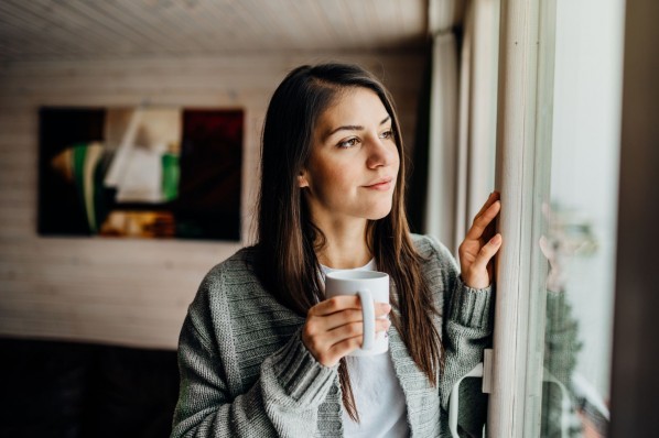 Eine etwa 25 jährige Frau steht an einem Fenster und schaut nach draußen. Sie hält eine Tasse in de Hand.