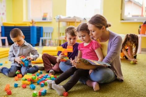 Kita-Erzieherin sitzt auf dem Boden und schaut mit 2 Kindern ein Buch an. Ein weiteres Kind sitzt daneben und spielt mit Bausteinen. 