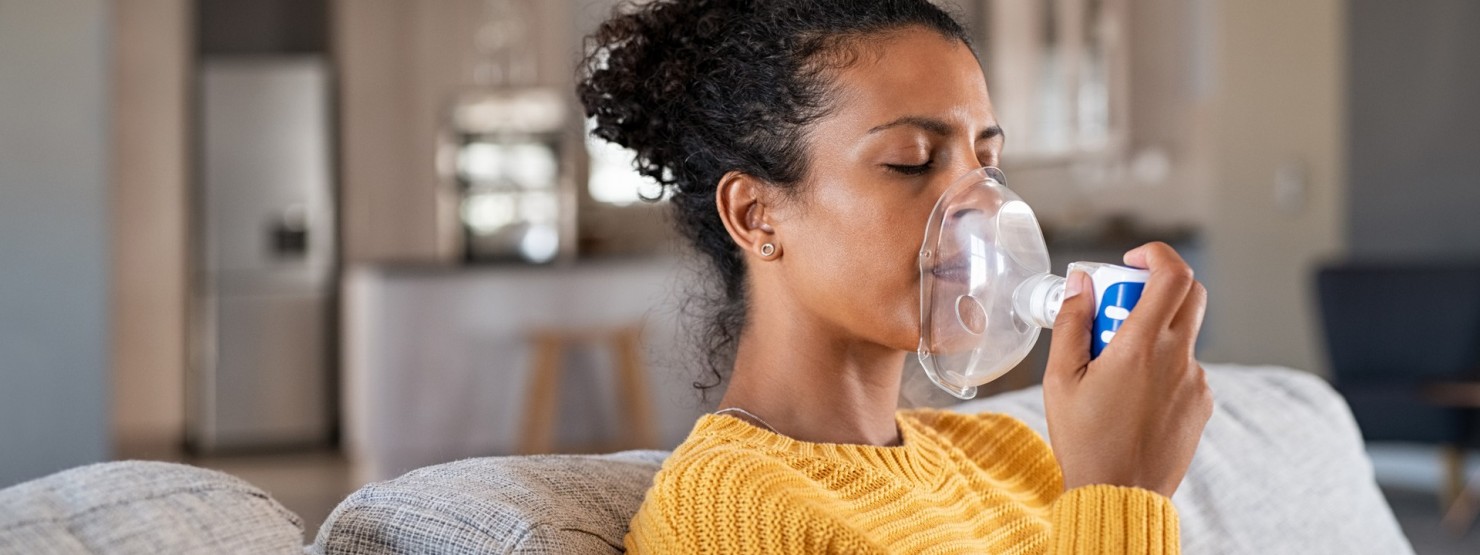 Eine Frau hat einen gelben Pullover an, sitzt auf einem Sofa und hält sich eine Inhalationsmaske an das Gesicht.