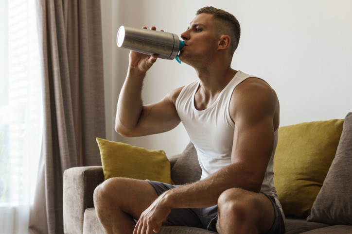Ein Kraftsportler sitzt auf dem Sofa und trinkt Kreatin aus einem Shaker.
