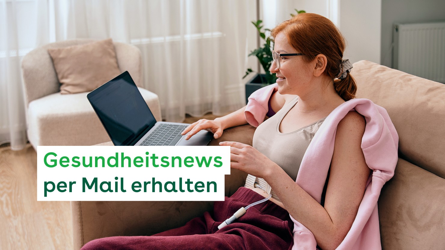 Eine etwa 25-jährige, rothaarige Frau sitzt in der Ecke ihres Sofas. Auf der Sofalehne steht ein Laptop, an dem sie arbeitet. Sie lächelt. Sie trägt ein bauchfreies Top, darunter ein Blutzuckermessgerät am Bauch.