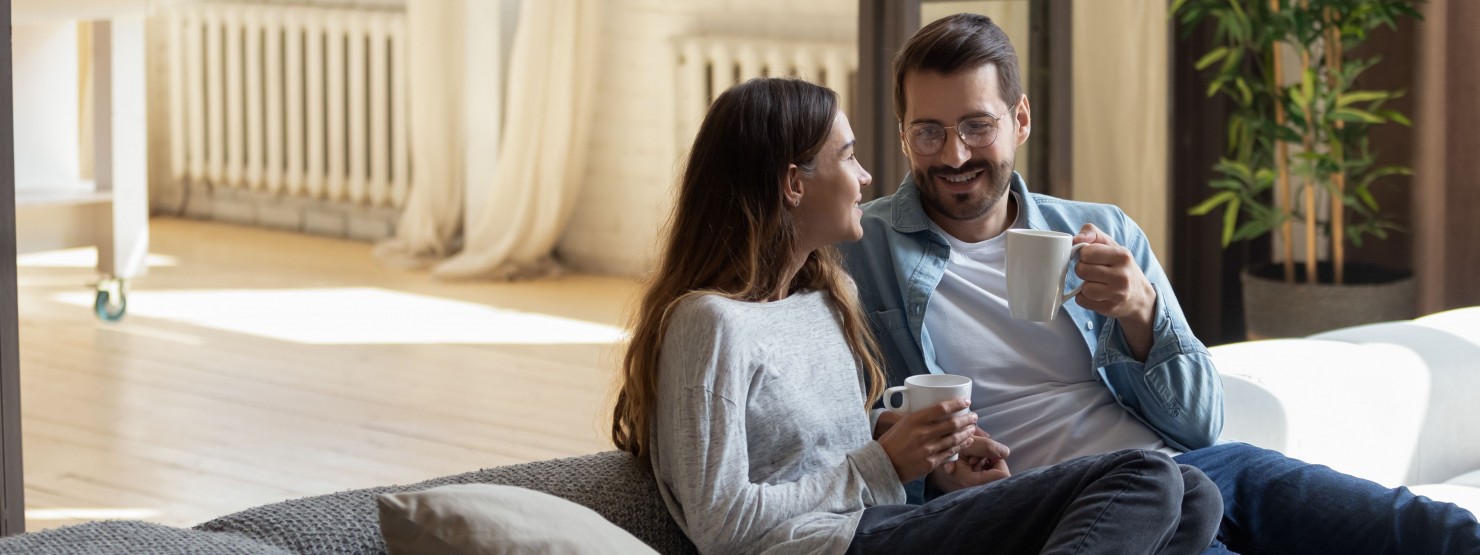 Ein Mann und eine Frau sitzen gemeinsam kuscheln auf einem Sofa. beide schauen sich lächelnd an und halten jeweils eine Tasse in der Hand.