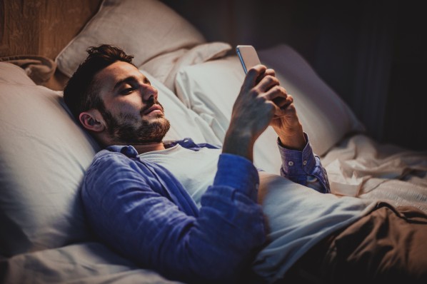 Ein etwa 30-jähriger Mann liegt im halbdunkel im bett und surft mit dem Smartphone im Internet