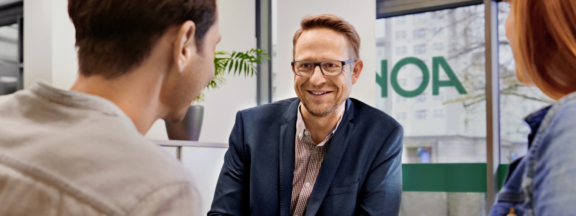 Ein etwa 40 jähriger Mann mit Brille und blauem Jakett unterhält sich mit 2 Personen. Sie befinden sich in einer AOK Filiale. An der Scheibe im Hintergrund ist das AOK-Logo angebracht.