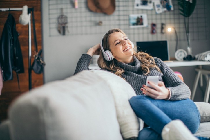 Eine etwa 30-jährige Frau liegt auf ihrer Couch und schaut auf sein Handy in der Hand. Er hört über Kopfhörer einen Podcast.