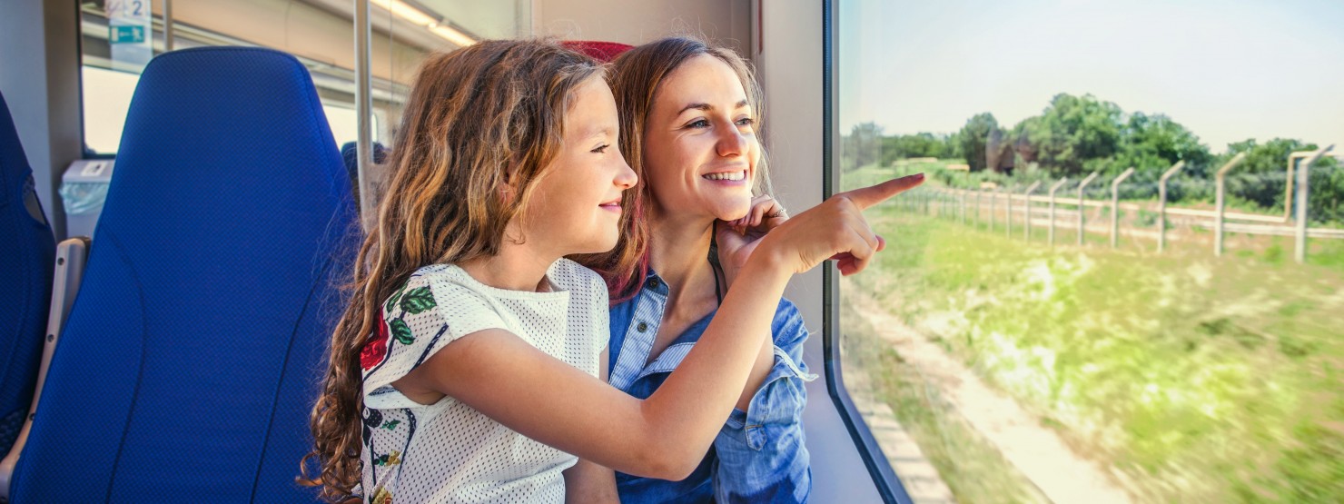 Mutter und Tochter sitzen zusammen im Zug und schauen lächelnd aus dem Fenster.