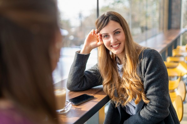 Eine etwa 25-jährige Frau sitzt lächelnd mit einer Freundin in einem Café.