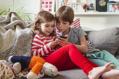 Zwei Kinder schauen gemeinsam auf ein Handy und sitzen auf einem Sofa.
