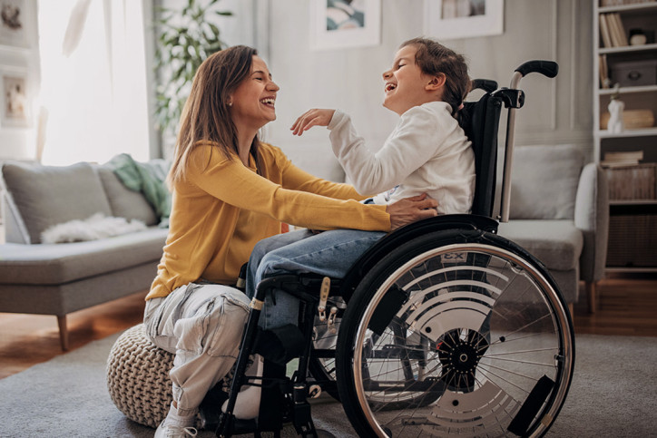 Eine etwa 40-jährige Frau in heller Jeans und gelbem Pullover sitzt auf einem Pouf vor ihrer etwa 10-jährigen Tochter, die im Rollstuhl sitzt. Sie kitzelt das Mädchen mit beiden Händen an den Körperseiten. Beide lachen ausgelassen. Sie befinden sich in einem hellen Wohnzimmer.