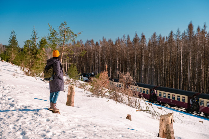 Frau mit gelber Mütze blickt beim Wandern auf die Bahngleise, auf denen die Brockenbahn gerade vorbeifährt