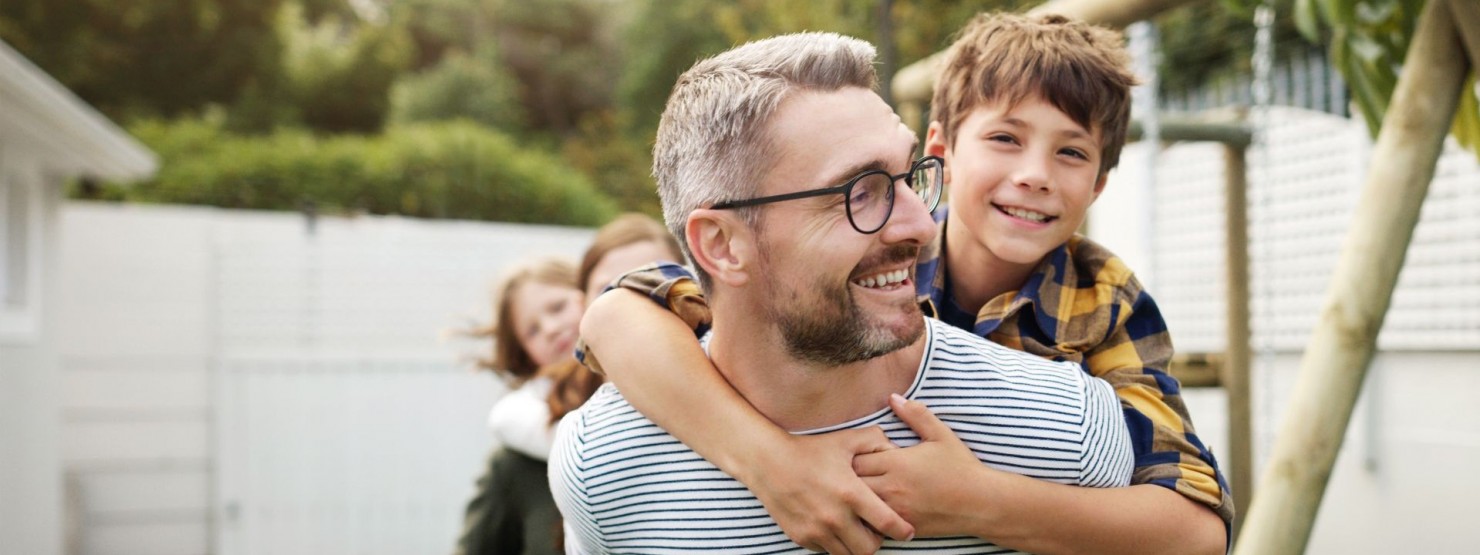 Ein Vater trägt seinen etwa 6 jährigen Sohn auf dem Rücken. Beide schauen sich an und lächeln.