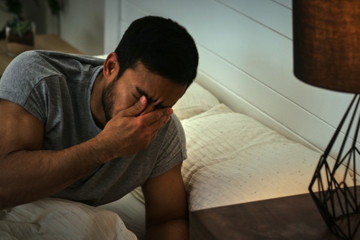 Ein junger Mann liegt im Bett und hält sich den Kopf, nachdem er Halluzinationen bei einer Schlaflähmung durchlitten hat.
