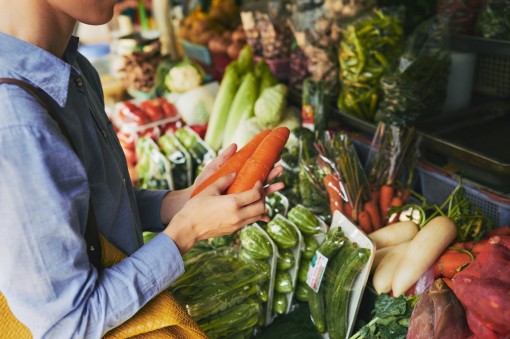 Eine etwa 30-jährige Person steht vor einem Gemüseregal und hält zwei frische Möhren in den Händen.