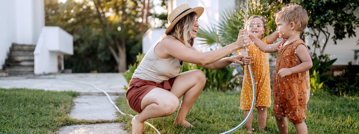 Junge Mutter ist mit ihren zwei Kindern im heimischen Garten. Sie besprüht die Kinder mit Wasser aus dem Gartenschlauch.