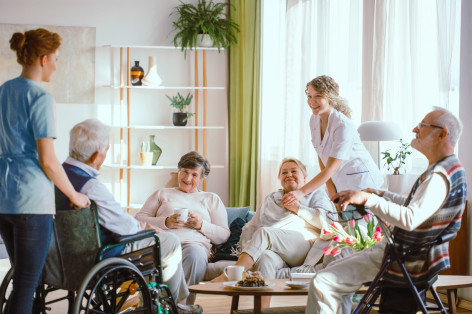 Vier Senioren sind zusammen in einem Wohnraum, erzählen und lächeln sich an. Zwei Pflegerinnen stehen bei und lächeln ebenfalls.