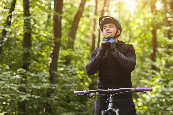 Ein etwa 30-jähriger Mann befestigt seinen Fahrradhelm. Er steht mit seinem Mountainbike in einem Wald.