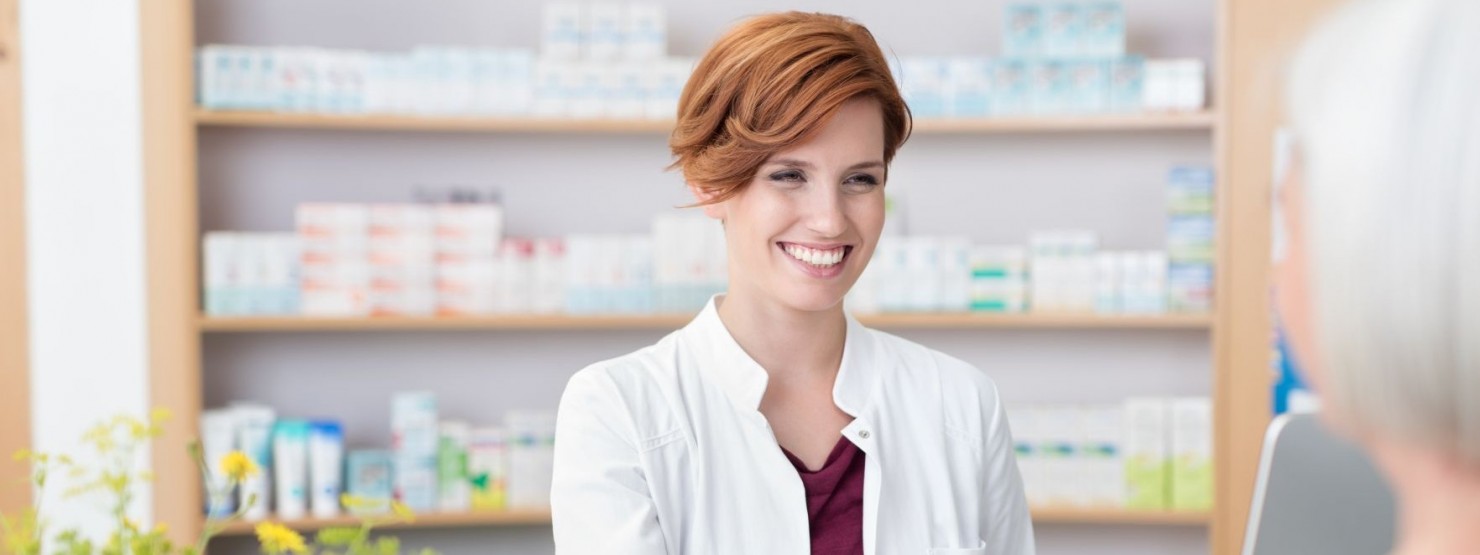 Eine Apothekerin gibt einer Frau ein Medikament über den Tresen aus. Im Hintergrund steht ein regal mit vielen weiteren Arzneimitteln.