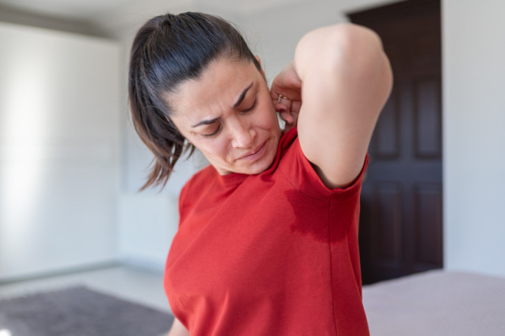 Eine Frau hebt ihren linken Arm. An ihrem T-Shirt zeichnet sich unter der Achsel ein Schweißfleck ab.