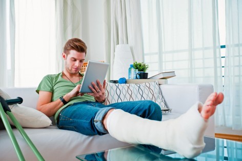 Ein junger Mann sitzt mit gebrochenen Bein auf dem Sofa und hat ein Tablet in der Hand.
