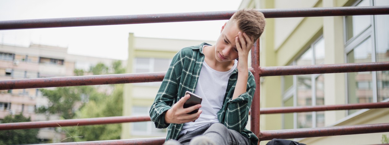 Ein ca. 14-jähriger Junge sitzt vor der schule auf einer Stufe und scha ut traurig auf sein Smartphone