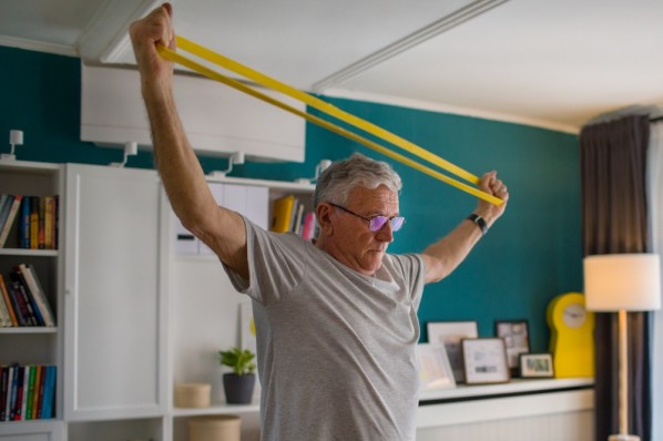 Mann Anfang 60 macht Übungen mt einem gelben Fitnessband in seinem Wohnzimmer