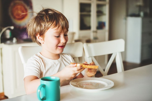 Ein etwa 5 jähriger Junge sitzt am Küchentisch und isst ein Marmeladentoast.