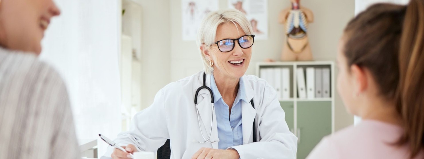 Eine etwa 50 jährige-Ärztin spricht mit einer Patientin und deren Mutter im Behandlungszimmer. Alle drei sitzen am Tisch.