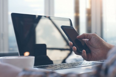 Eine Person hält in der rechten Hand ein Handy und sitzt vor einem Laptop.