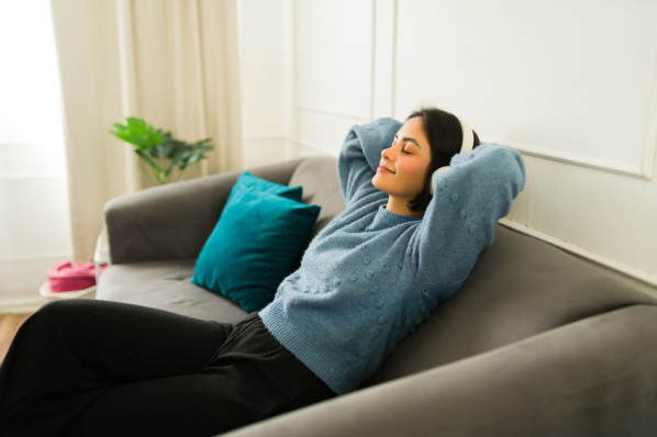Friedliche Frau, die sich auf einer weichen Couch entspannt, Musik über Kopfhörer hört, die Augen geschlossen, die Hände hinter dem Kopf ruhend