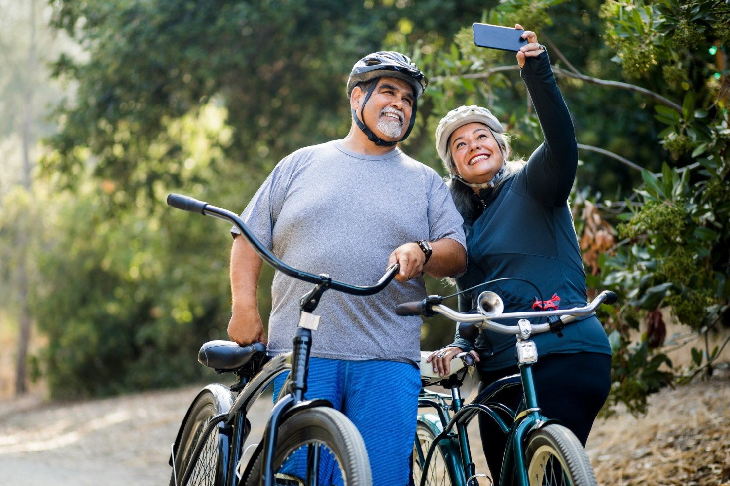 Ein Paar, Anfang 70, steht mit ihren Fahrrädern draußen im Grünen und macht ein Selfie. Sie tragen beide Fahrradhelme und lächeln in das Smartphone, das die Frau hält. Beide sind überwichtig. Sie strahlen Aktivität und Energie aus.