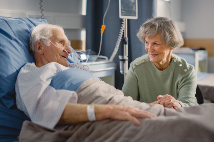 Ein älterer Patient liegt in einem Krankenhausbett und hat Besuch von einer Angehörigen. Er nimmt den Qualitätsvertrag zum postoperativen Delir in Anspruch.