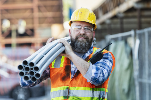 Ein Bauarbeiter läuft über eine Baustelle und trägt auf der Schulter mehrere Rohre.
