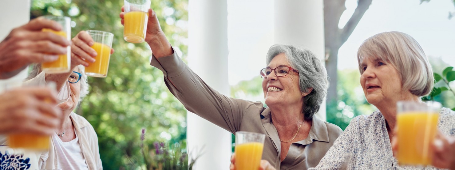 Eine Gruppe von etwa 70-jährigen Frauen sitzt im Freien. Sie prosten sich mit Orangesaft zu.