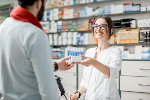 Ein Mann steht in der Apotheke und wird von einer Apothekerin beraten. Sie reicht ihm eine Medikamentenverpackung über den Thresen und lächelt dabei.