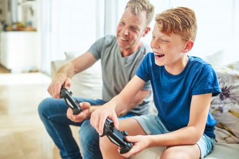 Ein Junge mit blauen Tshirt und Zahnspange sitzt mit einem erwachsenen Mann auf dem Sofa und spielt auf der Spielkonsole. 