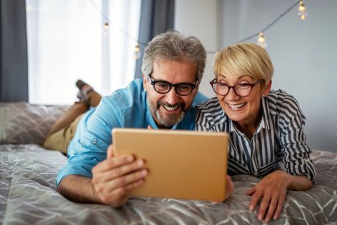 Ein etwa 55 jähriges Pärchen liegt auf einem Bett. Sie schauen gemeinsam auf ein Tablet und lachen.