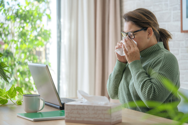Frau in grünem Pullover niest im Homeoffice in ein Taschentuch