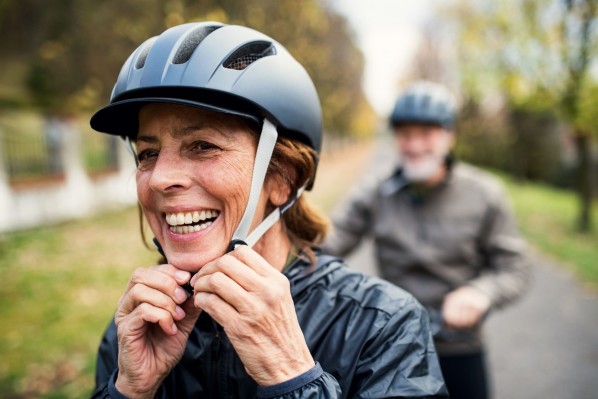 Eine etwa 60 jährige Frau fährt Fahrrad und trägt einen Helm. Dabei lächelt sie ausgiebig. Im Hintergrund befindet sich ihr Partner, der ebenfalls Fahrradfährt.