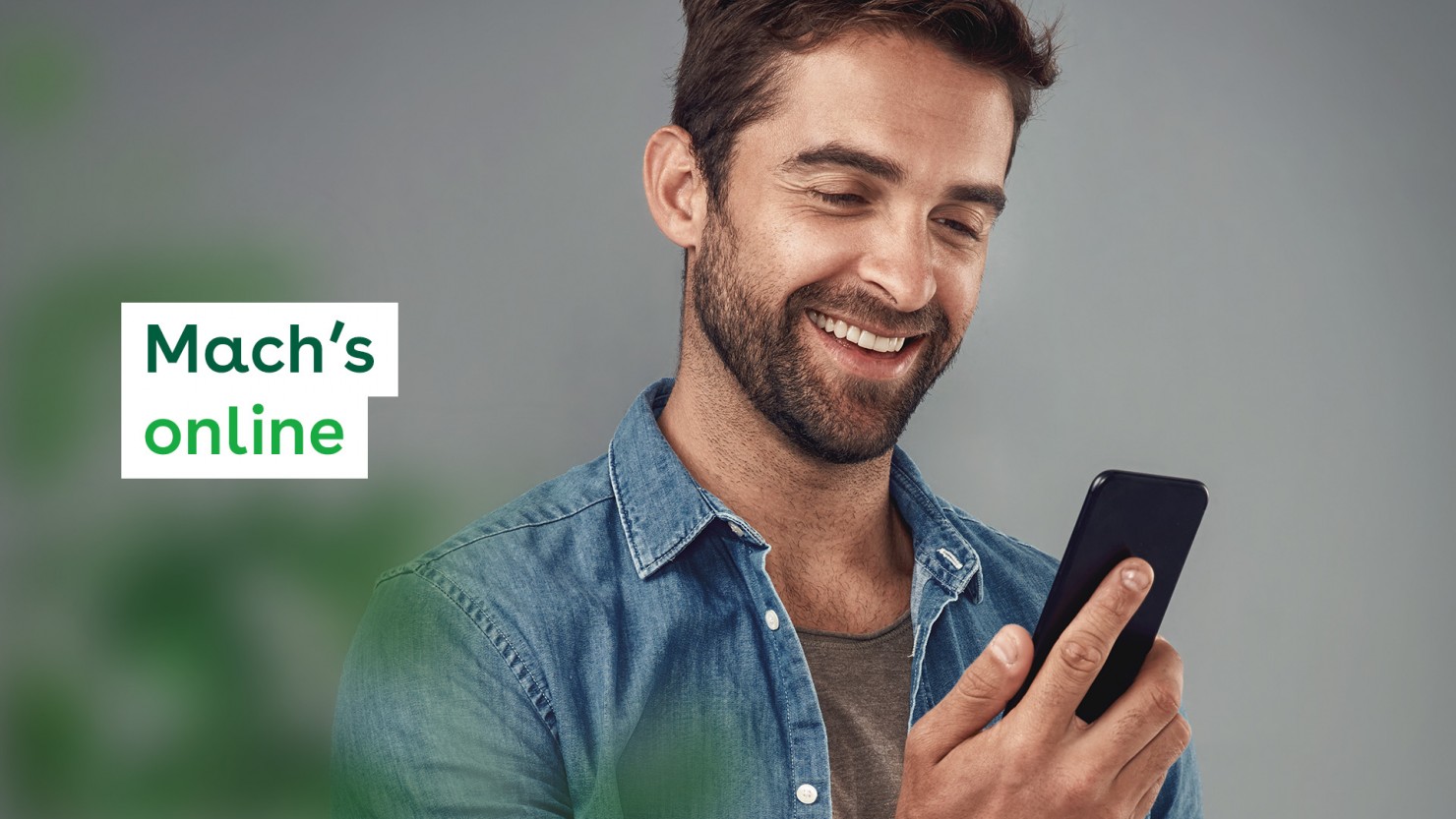 Ein Mann in einem Jeanshemd, etwa 40 Jahre, schaut lächelnd auf sein Smartphone, das er in der Hand hält.