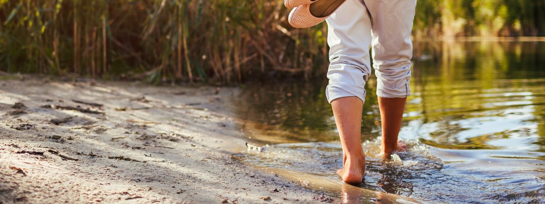 Eine etwa 55 jährige Frau steht unter Bäumen in der Natur und streckt ihre Arme in die Luft aus. Dabei schaut sie nach oben und lächelt.