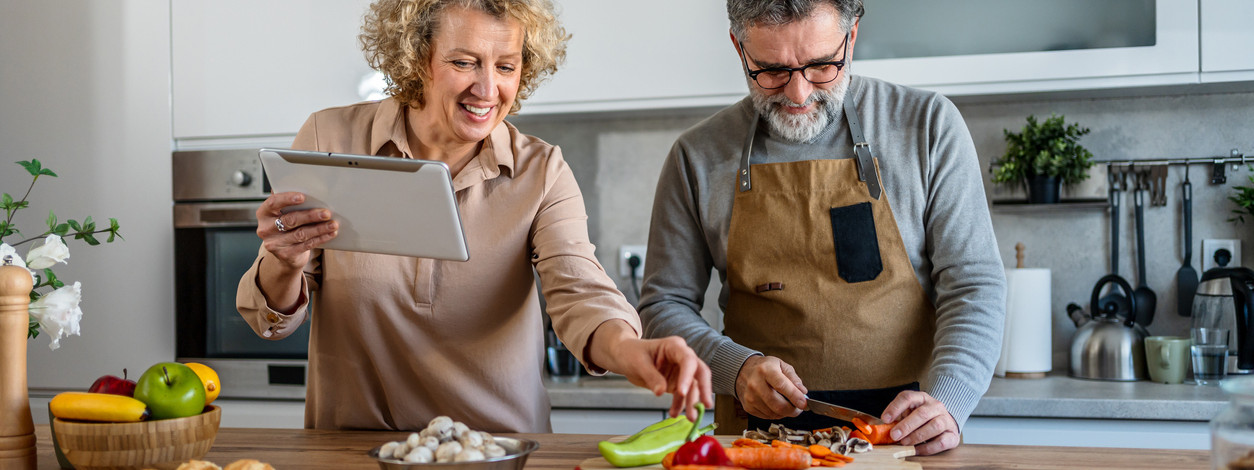 Glückliches reifes Paar, das zusammen nach einem Rezept auf einem digitalen Tablet kocht