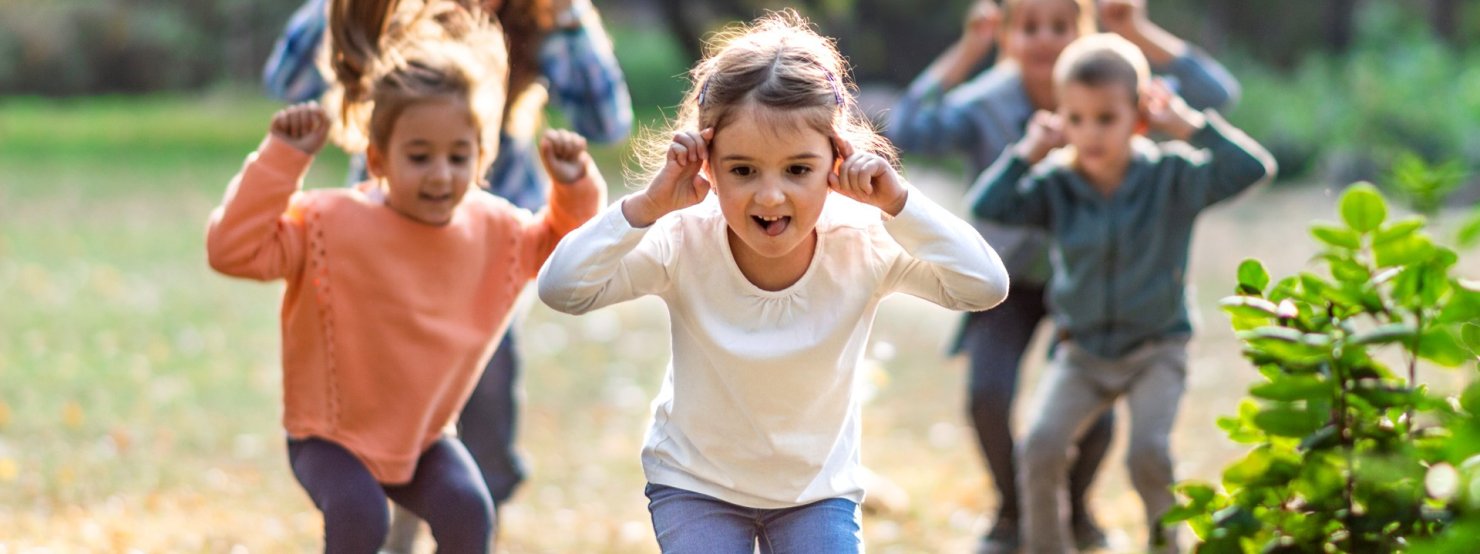 Kinder spielen im Freien, greifen sich an die Ohren und hüpfen dabei.