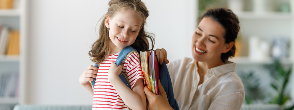 Ein Grundschulkind trägt einen Schulranzen. Die Mutter packt Bücher und Hefte in den Ranzen.