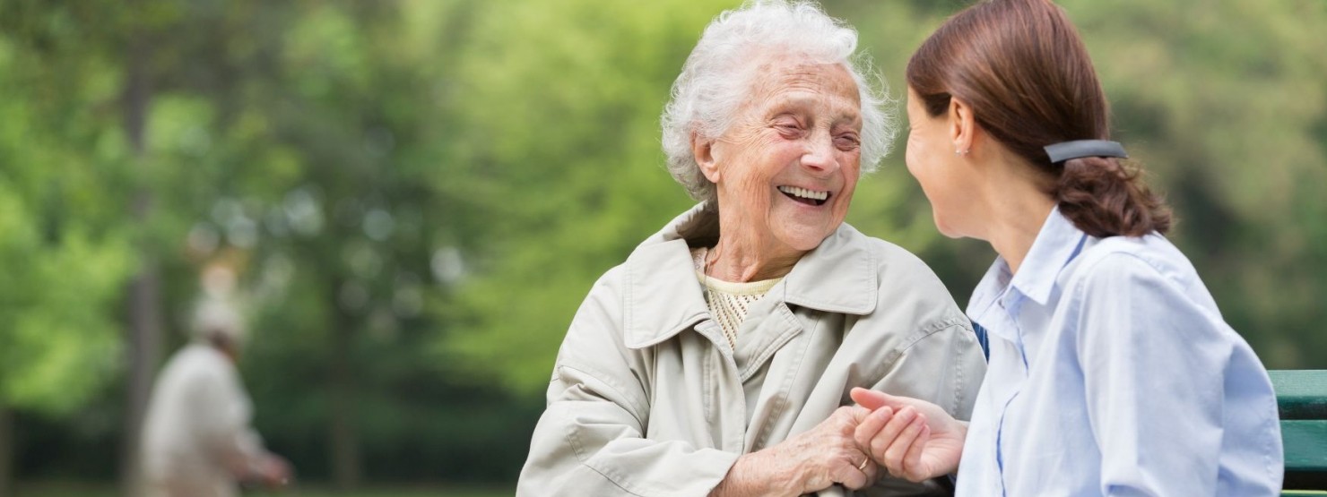 Eine Seniorin sitzt mit einer Frau auf einer Parkbank. Beide reichen sich die Hände und lächeln sich an.