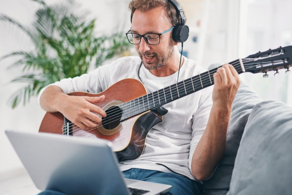 Ein etwa 40-jähriger Mann spielt Gitarre auf dem Sofa, während ein Laptop auf seinem Schoß liegt.