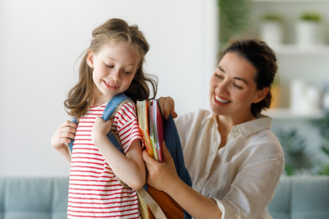 Ein Grundschulkind trägt einen Schulranzen. Die Mutter packt Bücher und Hefte in den Ranzen.